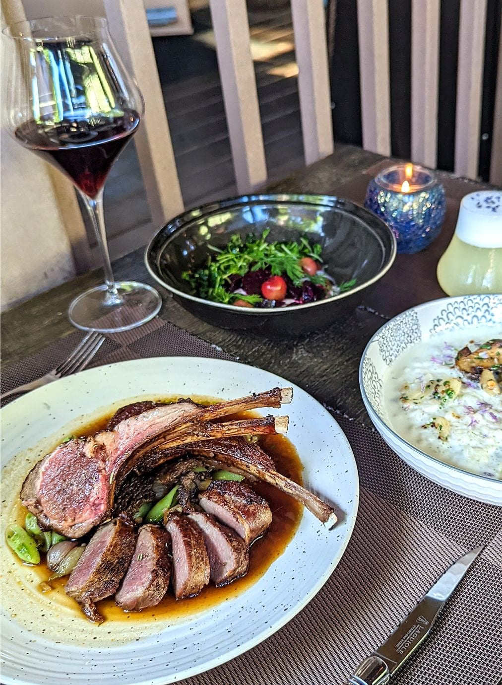 Sliced rack of lamb with jus, a side salad, risotto and a glass of red wine on a patio table