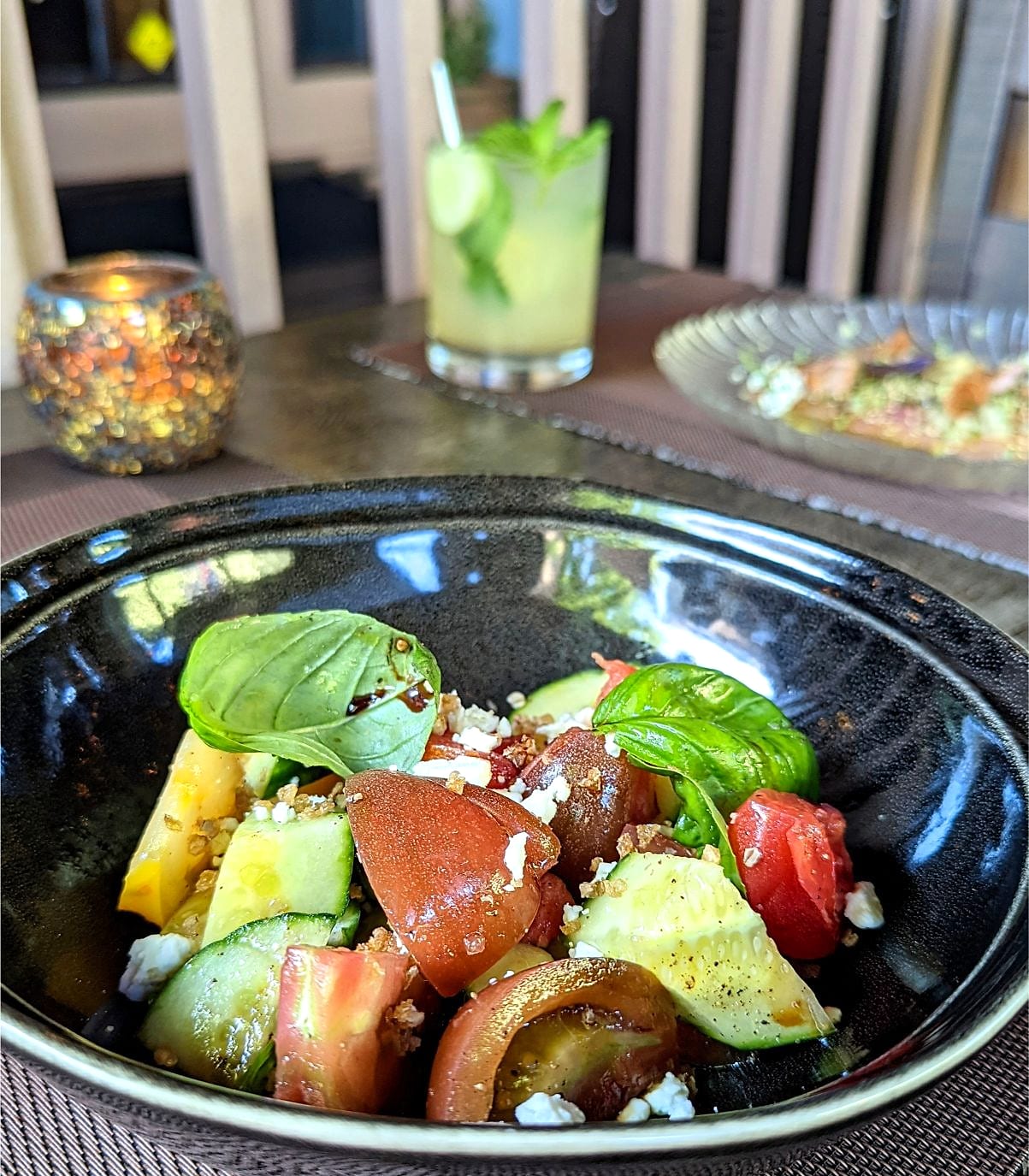 Heirloom tomato and cucumber salad with basil and cheese in a dark bowl, a cocktail behind
