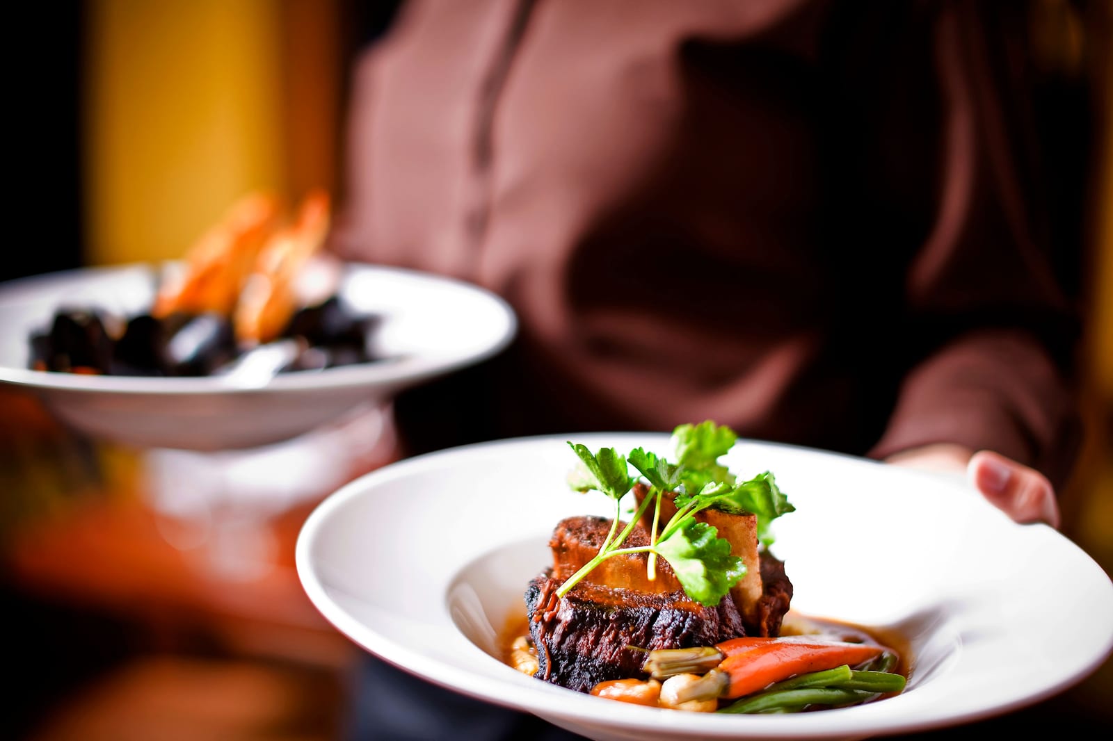 A server carrying a plated braised short rib with vegetables and a bowl of mussels