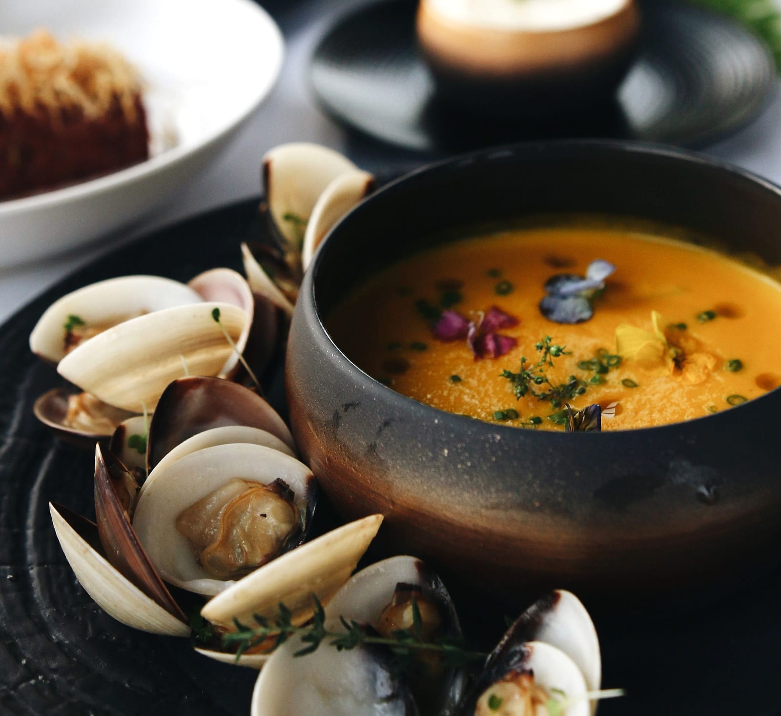 A bowl of bisque garnished with herbs and flowers, ringed with steamed clams and mussels