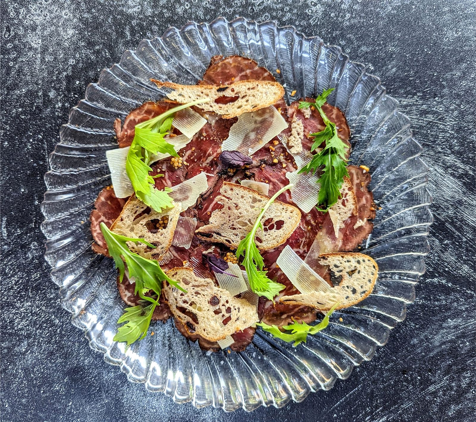 Beef carpaccio with crostini, shaved cheese and herbs on a fluted glass plate